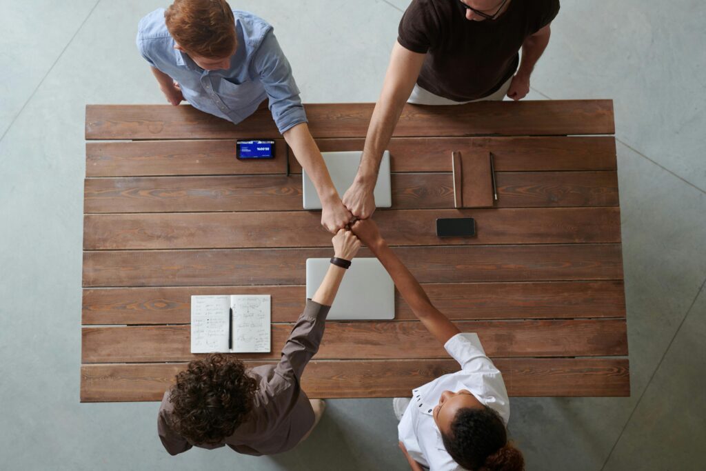 pexels-photo-3184430-3184430 A diverse group of professionals engaging in a teamwork celebration with a fist bump over a wooden table indoors.